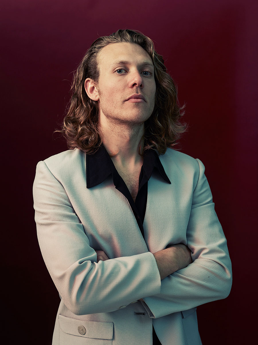 Studio portrait of musician, Jimmy Morris stood against a dark red background.